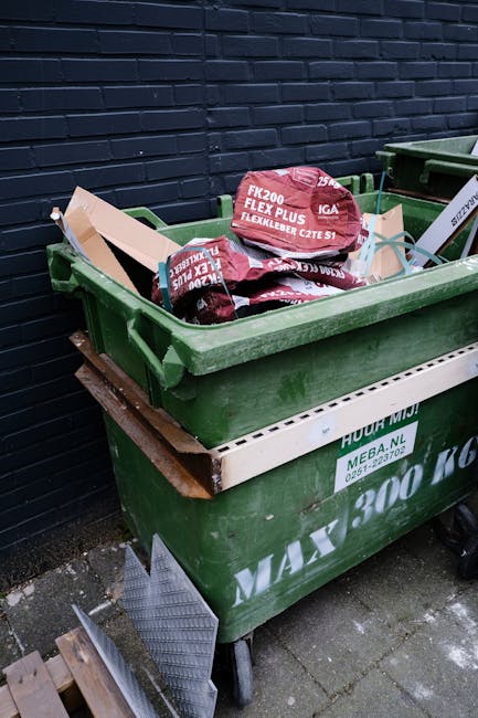 A large green plastic waste skip positioned on a paved surface next to a dark brick wall, filled with several bags of red and black flexible garden netting or fencing material. The skip has a white la