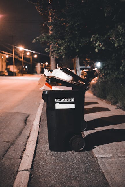 A large green plastic waste skip positioned on a paved surface next to a dark brick wall, filled with several bags of red and black flexible garden netting or fencing material. The skip has a white la